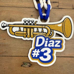 May include: A gold and white trumpet-shaped pendant with the name "Diaz" and the number "3" in blue and white. The pendant is attached to a blue and white chain. The background is a wooden surface.