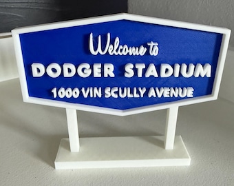 Puede incluir: Un letrero impreso en 3D que dice "Welcome to Dodger Stadium" en letras blancas sobre un fondo azul. El letrero incluye la dirección "1000 Vin Scully Avenue" y está sostenido por dos postes blancos y una base.