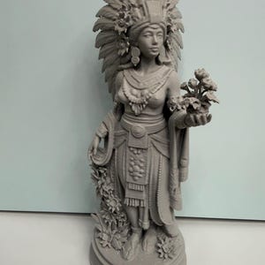 May include: A monochromatic gray 3D printed figurine of a figure wearing an elaborate feathered headdress and traditional layered garments. The figure holds a small bouquet of flowers in one hand and stands on a circular base with carved details.