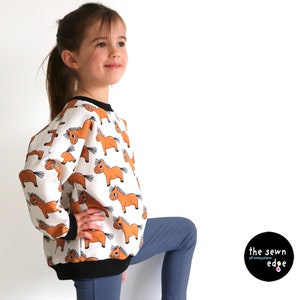 Pu&ograve; includere: Una felpa bianca con una stampa di cavalli marroni su tutta la superficie. La felpa ha un bordo a costine nero intorno al collo e ai polsini. Una giovane persona indossa la felpa e dei jeans blu.