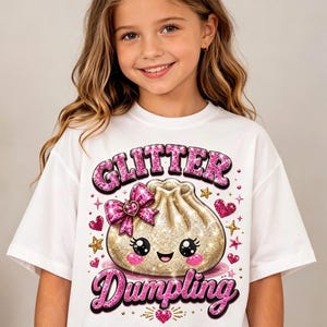 May include: A white t-shirt featuring a graphic of a gold glitter dumpling with a smiling face, pink cheeks, and a pink leopard print bow. The text GLITTER DUMPLING is printed in pink glittery letters, surrounded by small pink hearts and gold stars.