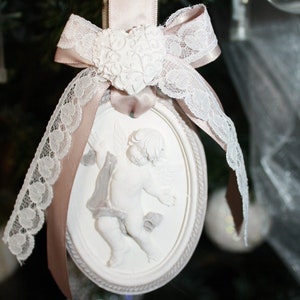 Op de afbeelding: Een wit porseleinen ornament met een cherub-ontwerp, versierd met een roze satijnen lint en witte kant. Het ornament is ovaalvormig en heeft een hartvormig ontwerp aan de bovenkant.