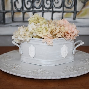 Peut inclure: Un pot en céramique blanche avec deux poignées, rempli d'un bouquet d'hortensias blancs et roses. Le pot est posé sur un plateau en céramique blanche avec un motif décoratif.