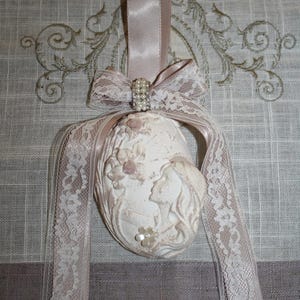 Può includere: Ornamento ovale color crema con un rilievo di due figure e accenti floreali. Presenta un nastro di raso rosa, un fiocco di pizzo con chiusura perlata e un delicato bordo in pizzo. Progettato per essere appeso.