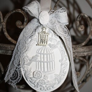 Può includere: Ciondolo in ceramica bianca con un design a forma di gabbia per uccelli e un piccolo uccello al suo interno. Il ciondolo è decorato con pizzo e un nastro di raso bianco.