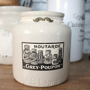 Può includere: Vaso in ceramica color crema con l'etichetta "Moutarde Grey-Poupon" e una grafica in bianco e nero di bottiglie di senape. Il vaso ha un bordo in pizzo e decorazioni metalliche. Altri vasi sono sullo sfondo.