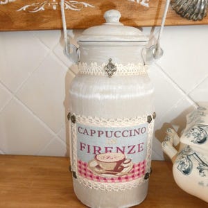 Op de afbeelding: Een witte keramische melkkan met een houten handvat en een kantversiering. De kan heeft een etiket met de tekst "Cappuccino in Firenze" met een afbeelding van een kopje cappuccino.