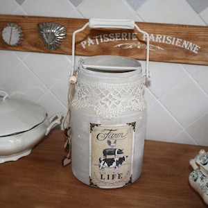 Può includere: Una latta di latte in metallo verniciata di bianco con un bordo in pizzo e un'etichetta in stile vintage che dice "Farm Life" con un gallo, un maiale e una mucca. La latta ha un manico bianco ed è appoggiata su una superficie di legno.