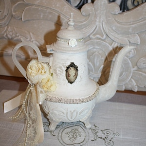 Può includere: Una teiera in ceramica bianca con un design floreale e un cammeo sul lato. La teiera è decorata con perle e pizzo e si trova su un tovaglia di lino bianco.