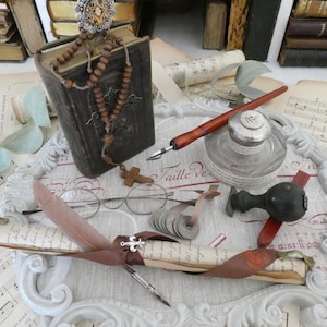 Peut inclure: Un livre vintage avec un chapelet sur le dessus, une plume, un encrier, un rouleau et un tampon sur une surface blanche avec une bordure décorative. Le livre a une couverture en cuir usée et le rouleau est attaché avec un ruban brun.