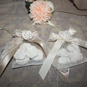 Può includere: Due piccole buste a rete trasparenti piene di caramelle bianche a forma di rosa. Ogni busta è adornata con un nastro di raso, finiture in pizzo e un abbellimento decorativo: una rosa di carta bianca e un cuore bianco. Un bouquet di fiori color pesca è sopra.