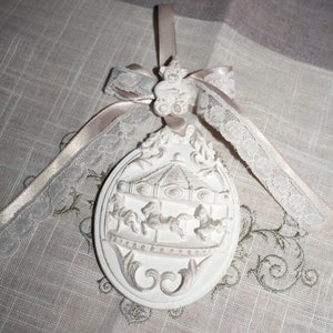 Peut inclure: Ornement ovale blanc avec un motif de manège, représentant des chevaux et un dessus décoratif. L'ornement est orné d'un ruban beige et d'une garniture en dentelle. Une petite figurine d'ours en peluche est au sommet.