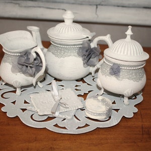 Puede incluir: Juego de té blanco con tetera, jarra de crema, azucarero y un mantel de encaje. El juego está decorado con encaje gris y flores.