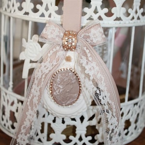 Puede incluir: Una jaula decorativa blanca con intrincados diseños calados. Una cinta rosa rubor y un lazo de encaje con un acento de joya adornan la parte superior. Un colgante de camafeo con un marco de oro rosa cuelga debajo.