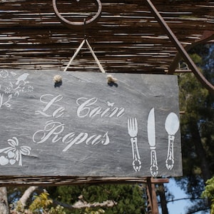 Peut inclure: Une enseigne en ardoise noire avec une inscription blanche qui dit "Le Coin Repas" et un graphique d'un couteau, d'une fourchette et d'une cuillère. L'enseigne est suspendue à une structure en bois.