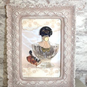 Peut inclure: Un flacon de parfum vintage dans un cadre décoratif. Le flacon est en verre transparent avec une étiquette dorée qui indique "Shalimar Guerlain Paris". Le cadre est de couleur rose clair avec un motif de dentelle.