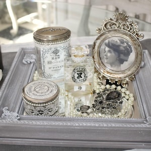 Peut inclure: Un plateau rectangulaire gris présente une collection d'objets vintage. On y trouve un flacon de parfum, un petit portrait encadré, deux boîtes décoratives, une broche et un collier de perles. Une rose blanche et une bougie sont également présentes.