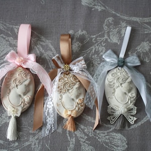Può includere: Tre ornamenti a cammeo ovali con nastri. Ognuno presenta un profilo femminile classico in rilievo, adornato con pizzo, fiocchi e nappe. I nastri sono rosa, beige e argento, creando un'estetica vintage delicata.