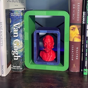 Puede incluir: Un busto rojo de un hombre con bigote se exhibe dentro de un marco azul, anidado dentro de un marco verde más grande. Los marcos están sobre un estante con libros, incluyendo títulos como "Van Gogh" y "Leonardo da Vinci".
