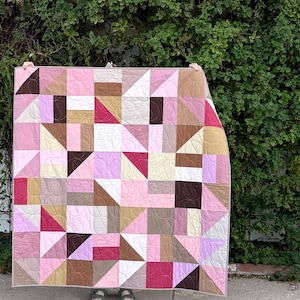 Op de afbeelding: Een geometrische quilt met een patchwork ontwerp van driehoeken en vierkanten in tinten roze, bruin en crème. De quilt wordt omhooggehouden tegen een achtergrond van weelderig groen gebladerte en een witte muur.
