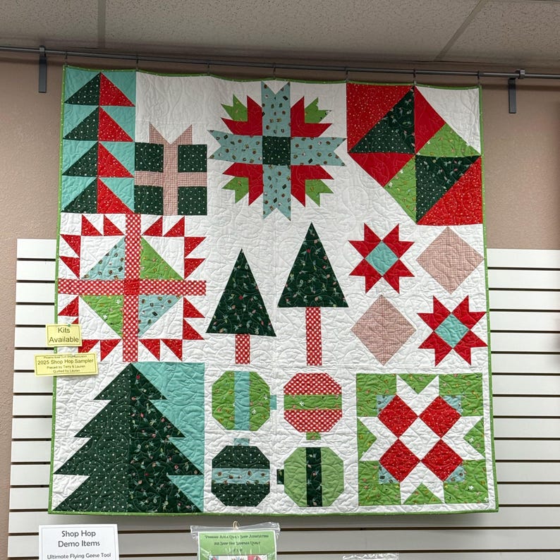 May include: A square patchwork quilt featuring a holiday theme with red, green, white, and light blue fabric blocks. The design includes patterns of Christmas trees, gift boxes, star shapes, and round ornaments arranged in a grid on a white background.
