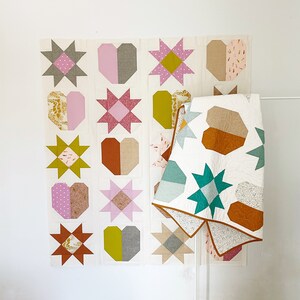 May include: A patchwork quilt with a design featuring hearts, stars, and geometric shapes in shades of pink, green, brown, and white. The quilt is made up of 16 individual squares, each with a different design. The quilt is hanging on a white wall.