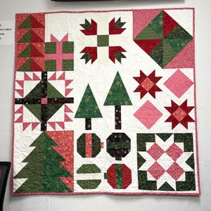 May include: A square patchwork quilt featuring a white background with various holiday-themed blocks including green Christmas trees, red and green gift boxes, star patterns, and circular ornaments in shades of red, green, pink, and black fabric.