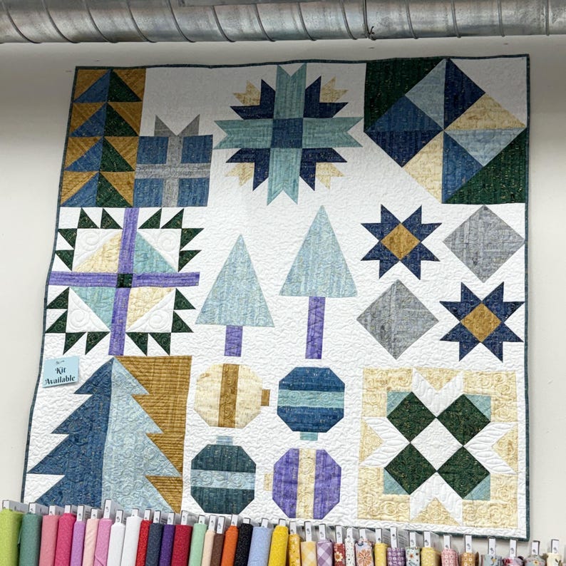 May include: A square quilt featuring a white background with various pieced blocks including geometric stars, stylized evergreen trees, and circular ornaments in shades of blue, green, yellow, and purple. A small blue tag reads Kit Available.