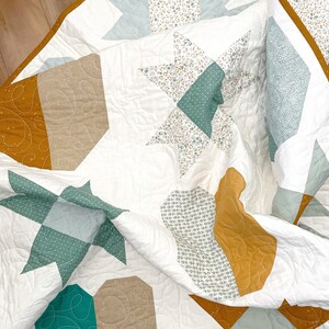 May include: A white quilt featuring a mix of patterns in shades of brown, green, and white. The quilt is composed of various geometric shapes, including stars, hexagons, and circles.