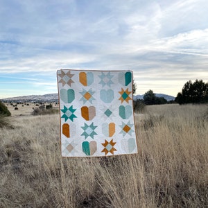 May include: A white quilt with a patchwork design featuring teal, blue, and orange geometric shapes. The quilt is hanging on a clothesline in a field of long grass.