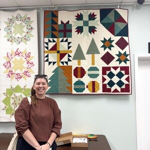 May include: A square patchwork quilt featuring a holiday-themed design with geometric patterns, stylized evergreen trees, gift boxes, and circular ornaments in shades of cream, forest green, deep red, mustard yellow, and teal, with visible quilting.