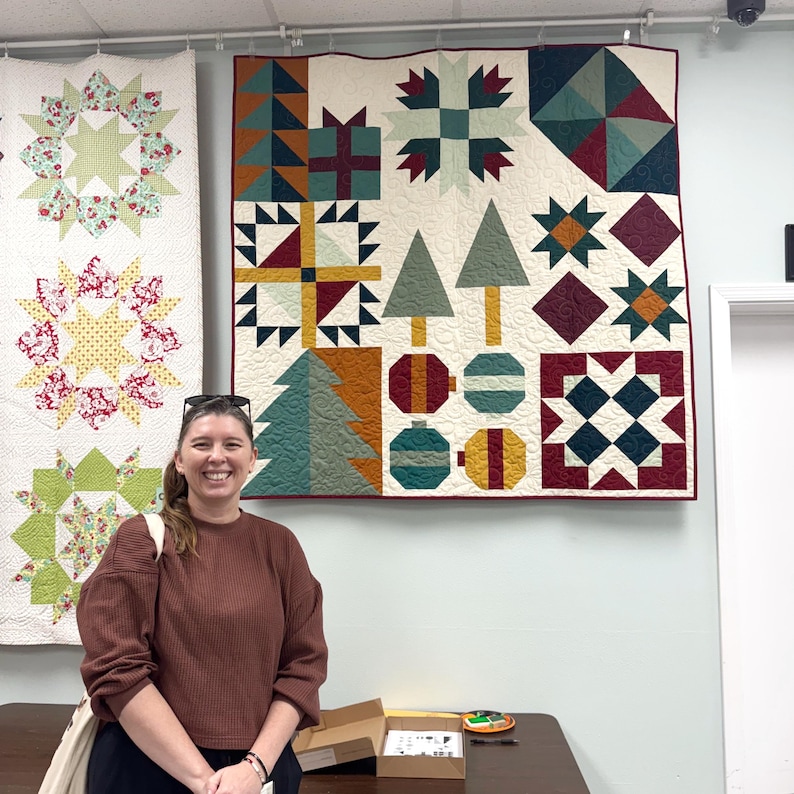 May include: A square patchwork quilt featuring a holiday-themed design with geometric patterns, stylized evergreen trees, gift boxes, and circular ornaments in shades of cream, forest green, deep red, mustard yellow, and teal, with visible quilting.