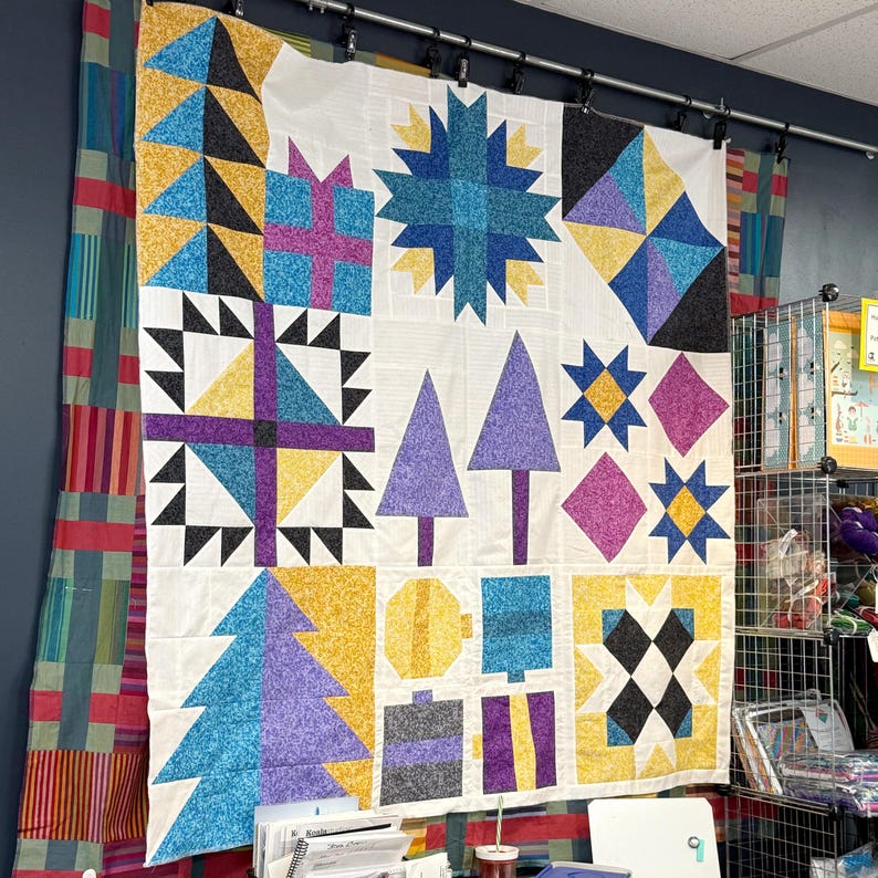 May include: A large square quilt featuring a patchwork design with various geometric shapes, including stars, trees, and gift boxes in vibrant shades of yellow, blue, purple, black, and white, displayed hanging against a dark wall.
