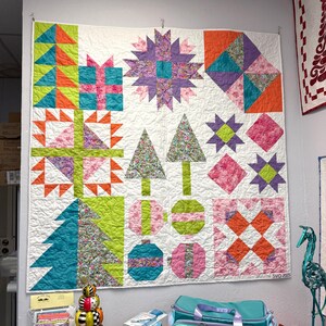 May include: A square patchwork quilt featuring a white background with various colorful blocks including geometric stars, stylized Christmas trees, gift boxes, and circular ornaments in shades of lime green, orange, purple, pink, and teal.