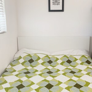 May include: A white bed with a green and white patterned quilt. The bed has a white headboard and is against a white wall.