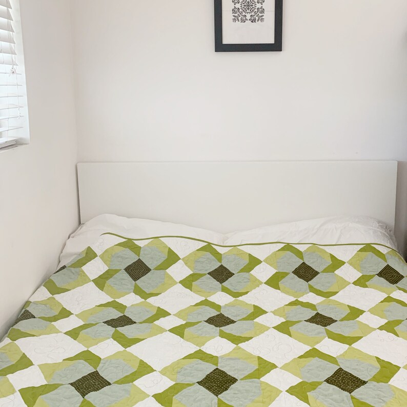May include: A white bed with a green and white patterned quilt. The bed has a white headboard and is against a white wall.