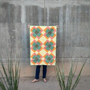 May include: A quilt with a geometric pattern of green, orange, and yellow squares. The quilt is being held up by a person, but the person's hands are not visible in the image.