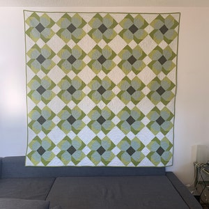 May include: A green and white quilt with a geometric pattern of overlapping squares and circles. The quilt is hanging on a wall above a gray couch.