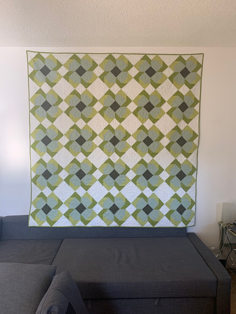 May include: A green and white quilt with a geometric pattern of overlapping squares and circles. The quilt is hanging on a wall above a gray couch.
