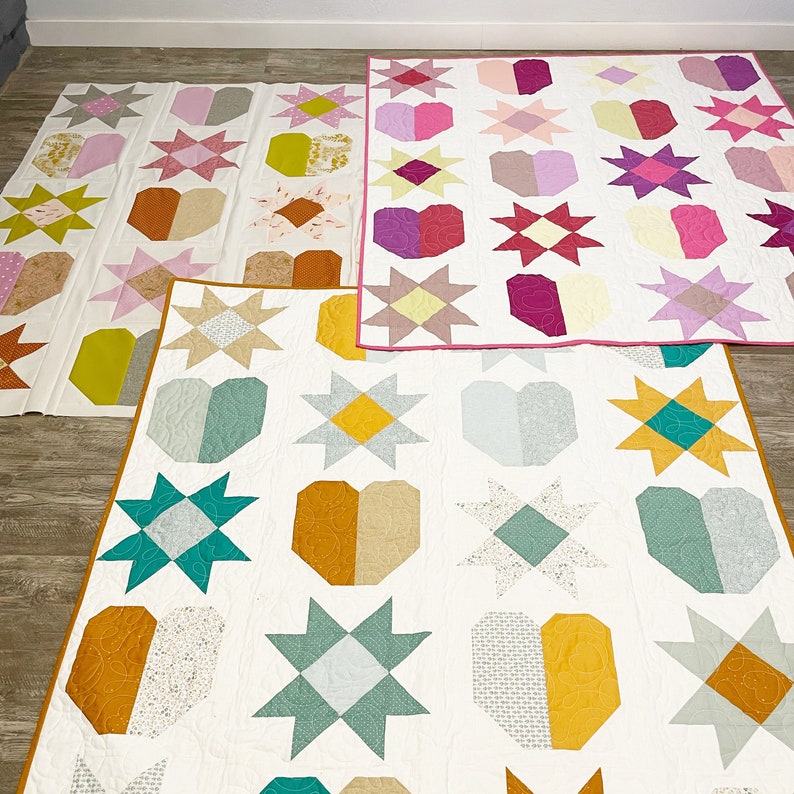 May include: Three quilt blocks featuring different patterns. The top block is pink, white and purple with a heart, star and flower design. The middle block is yellow, white and green with a heart, star and flower design. The bottom block is brown, white and green with a heart, star and flower design.