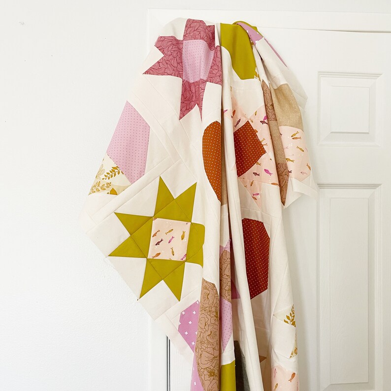 May include: A white quilt with a patchwork design featuring pink, orange, and yellow fabric squares. The quilt is made of different patterns and textures, including stars, fish, and geometric shapes.