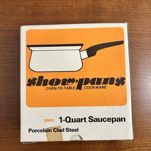 May include: A white cardboard box for a Show-Pans 1-Quart Saucepan. The box features an orange square with a black and white illustration of a saucepan, the text "show-pans OVEN-TO-TABLE COOKWARE", "2001 1-Quart Saucepan", and "Porcelain Clad Steel".