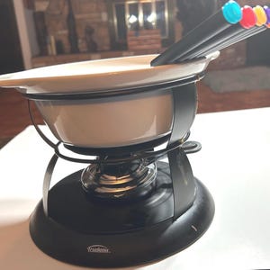 May include: A fondue set with a cream-colored ceramic pot, black metal stand, and burner. The set includes black fondue forks with colorful tips. The base of the stand has the brand name "Trudeau" printed on it.