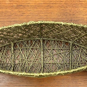 以下が含まれることがあります： ユニークな細長い楕円形をした、オリーブグリーンの織りバスケット。バスケットは、絡み合った繊維のネットワークで構成されており、開放的で風通しの良いデザインになっています。バスケットの縁はきれいに仕上げられています。