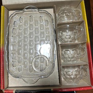 Puede incluir: Un juego de aperitivo de cristal transparente que consta de una bandeja rectangular con una hendidura circular y cuatro tazas a juego. La bandeja y las tazas presentan un patrón ovalado en relieve. Los artículos están en una caja.