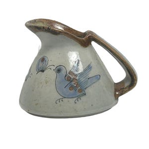 May include: A ceramic creamer with a speckled off-white glaze and a brown rim and handle. The body features a hand-painted light blue bird with brown circular accents on its wing and a small blue butterfly motif on the side of the vessel.