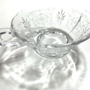 Pode incluir: Chávena de chá de vidro transparente com uma pega e um padrão floral decorativo gravado na superfície. A chávena tem uma borda ondulada e assenta numa superfície branca, projetando uma sombra.