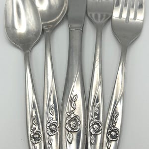 Op de afbeelding: Een set zilverkleurig bestek, bestaande uit twee lepels, een mes en twee vorken. Elk stuk is versierd met een bloemenroosontwerp op het handvat. Het bestek is gerangschikt op een wit oppervlak.