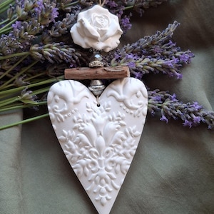 May include: A white heart-shaped ornament featuring an intricate embossed floral pattern, topped with a small wooden stick, a silver-toned bead, and a sculpted white rose. A thin twine loop is attached at the top for hanging purposes.
