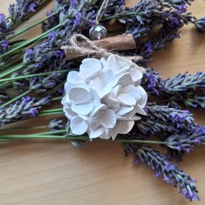 Peut inclure: Une décoration en forme de fleur d'hortensia en argile blanche, agrémentée d'un petit bâton de cannelle brun et d'une perle à facettes transparente, suspendue par une ficelle, posée sur un bouquet de tiges de lavande séchée violette.